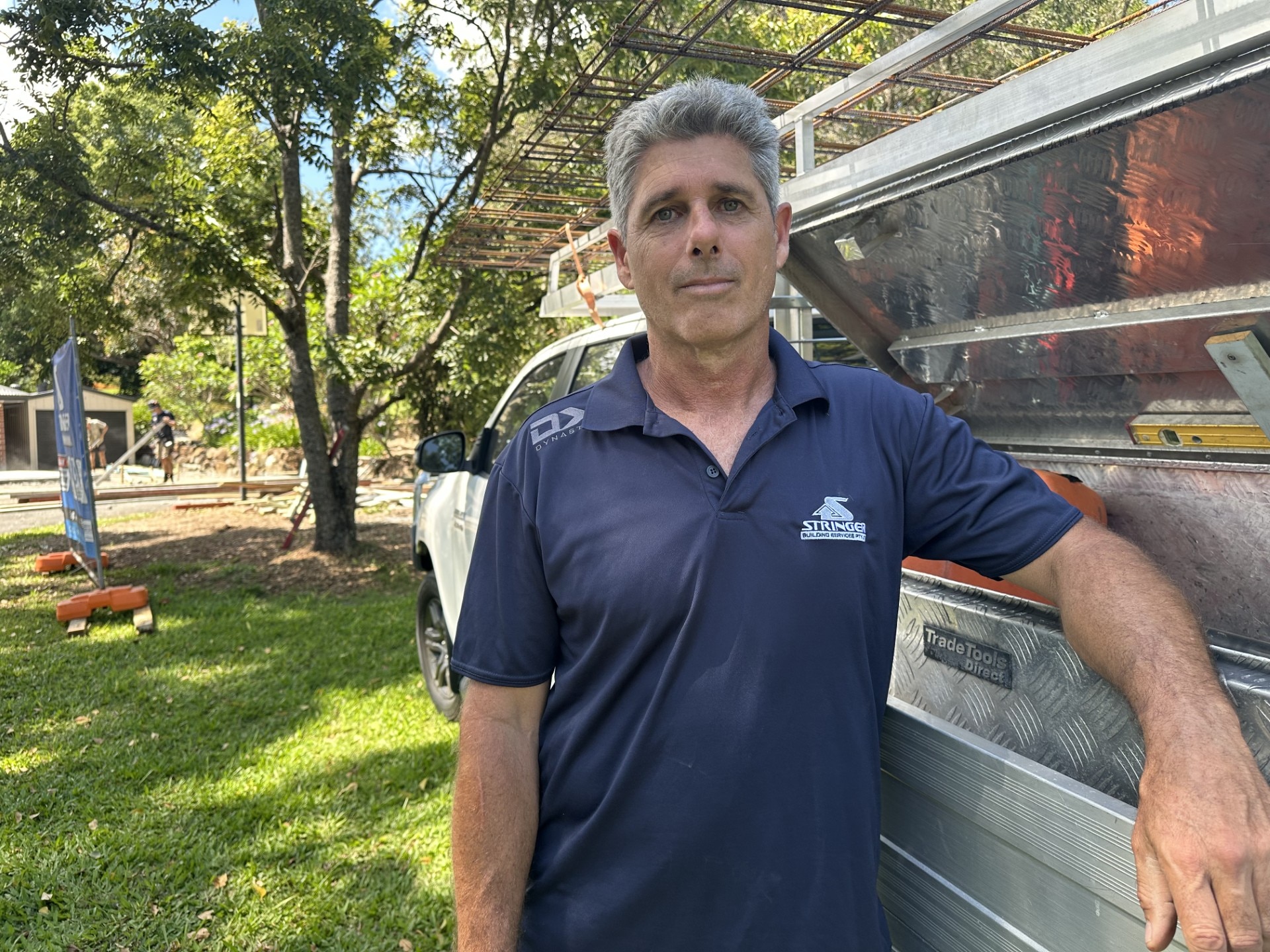 Un hombre de pelo gris que lleva un polo con el logo se apoya en una camioneta estacionada en el césped.