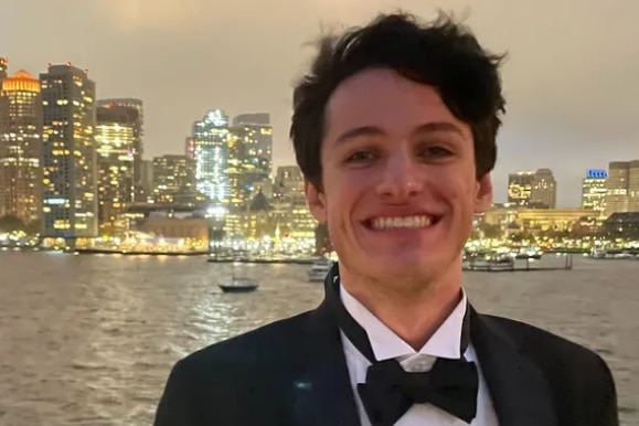 An image of a young man smiling into a camera while wearing a tuxedo, city lights beyond the river.