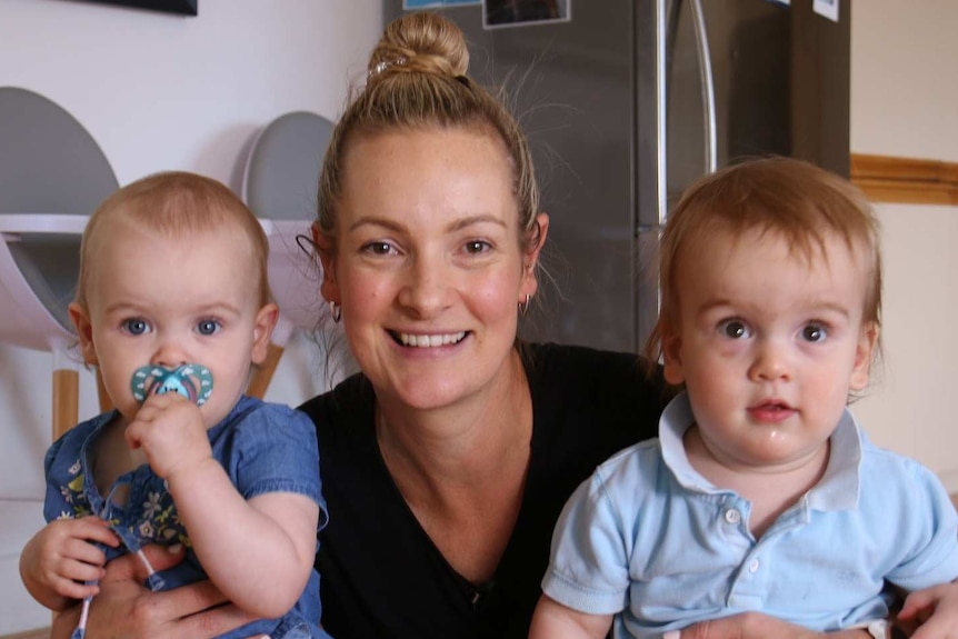 A woman smiles at the camera and squats down in a kitchen with her arms around her two young children.