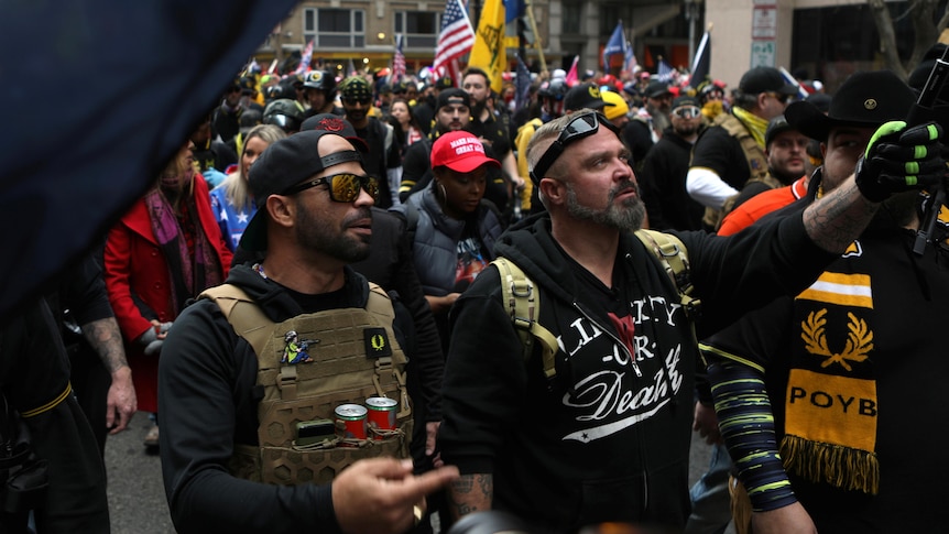 Two men decked out in tactical gear walk through a crowd with American flags and yellow and black designs visible