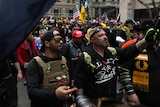 Two men decked out in tactical gear walk through a crowd with American flags and yellow and black designs visible