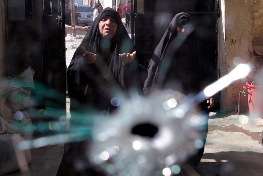 An elderly woman with a pained look on her face and wearing a head scarf holds out hands. View through bullet shot through glass