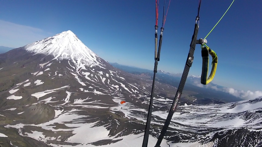 Set amongst snow-capped mountains, a man flies below a parachute in the mid-ground.