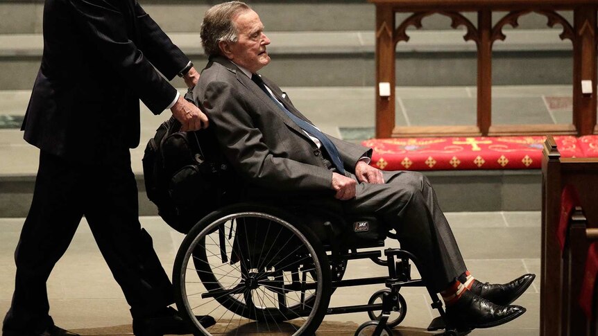 A middle-aged white man in a suit pushes an elderly white man wearing a suit seated in a wheelchair