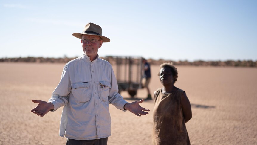 white man with wide-brimmed hat arms outstretched standing new to aboriginal woman in desert