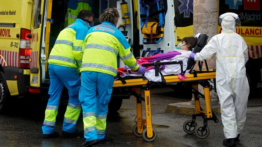 Healthcare workers in masks and one in a full biohazard suit push a patient on a stretcher into a yellow ambulance.