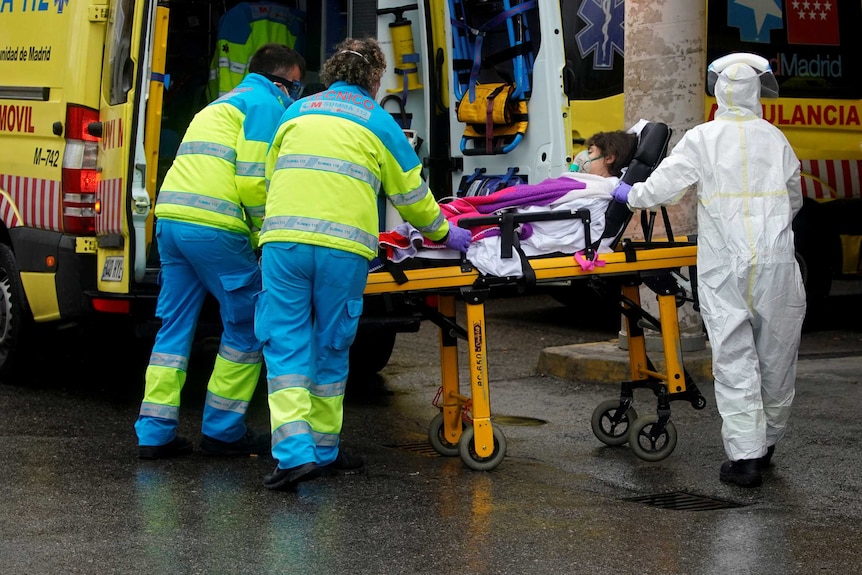 Healthcare workers in masks and one in a full biohazard suit push a patient on a stretcher into a yellow ambulance.