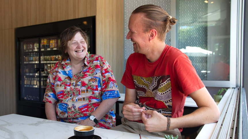 A smiling woman in a colourful shirt looking at a man in a red shirt who is smiling back at here, both sitting in a cafe.