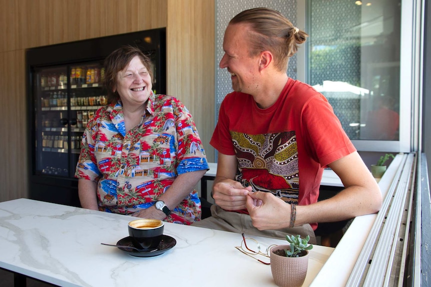 A smiling woman in a colourful shirt looking at a man in a red shirt who is smiling back at here, both sitting in a cafe.