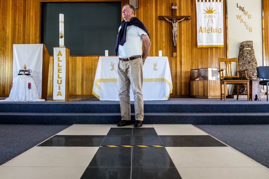Catholic Priest, Father Mick Lowcock, stands casually in his church, his jumper tied around his shoulders 