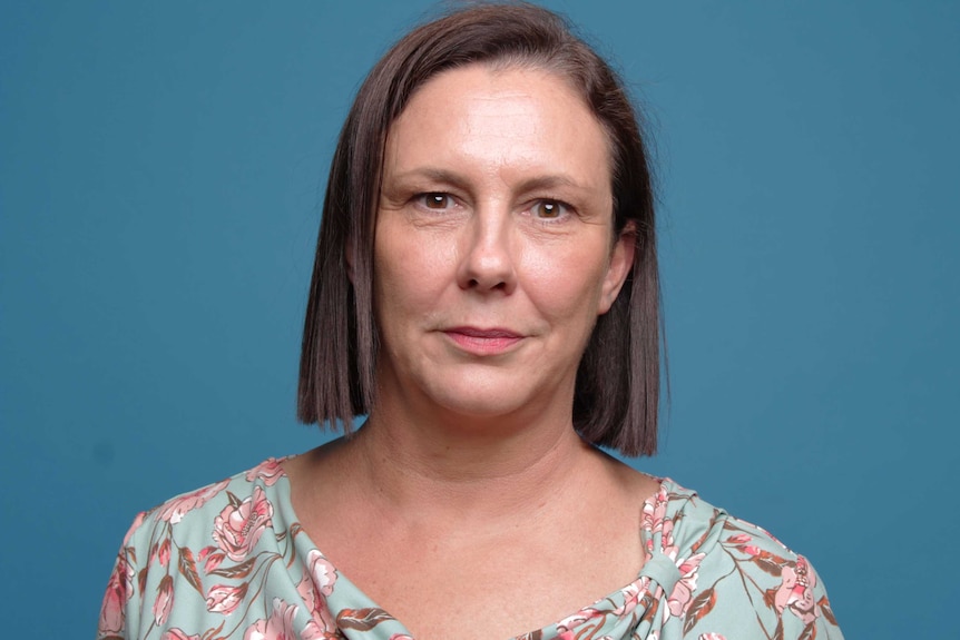 Woman with floral top and neutral expression, before a blue background.