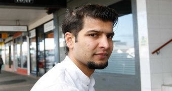 A man with a goatee and a white shirt stands  on a footpath and  looks into the camera.