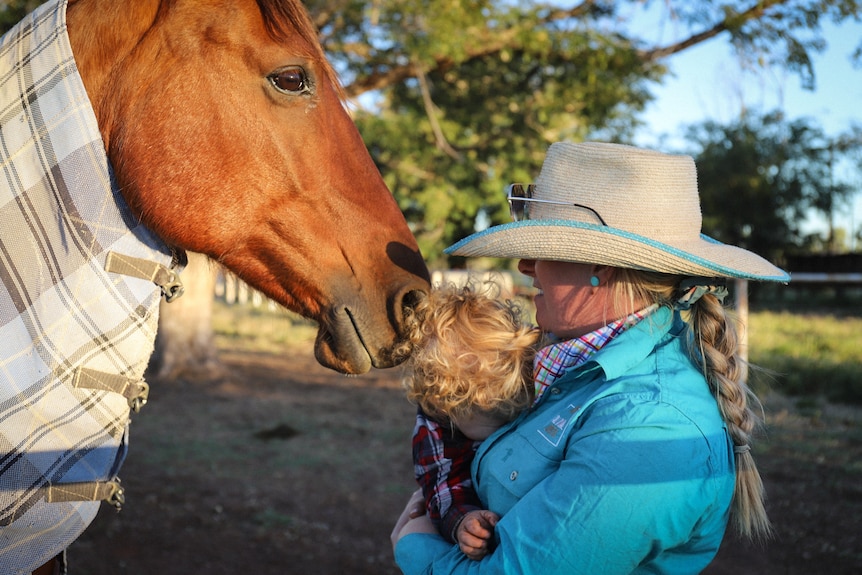 a woman in bush hat and jade shirt holds a baby in front of her as they get close to a horse's face/head