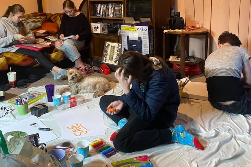Teenagers sit on floor of living room, writing cardboard slogans to rally to keep aged care home open