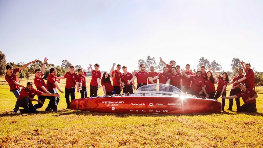 24 young adults in red shirts and jeans in a humorous pose in a paddock on a sunny day presenting a shiny red solar car.