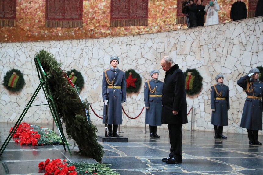 Putin bows his head in front of an enormous wreath in a stone memorial hall. Guards stand by, but none hold guns