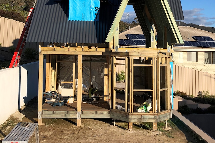 The wooden frame of what looks like a small house, but is in fact a toy for children, stands in a backyard.