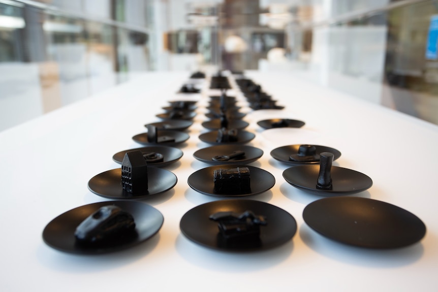 Three rows of black cast glass small sculptures of dishes with most holding different objects like cars, buildings and a boot