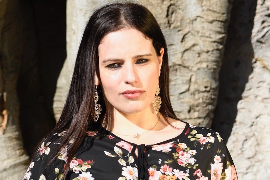 Woman in a black floral blouse and chandelier earrings looks seriously at the camera as her dark hair falls over her shoulders