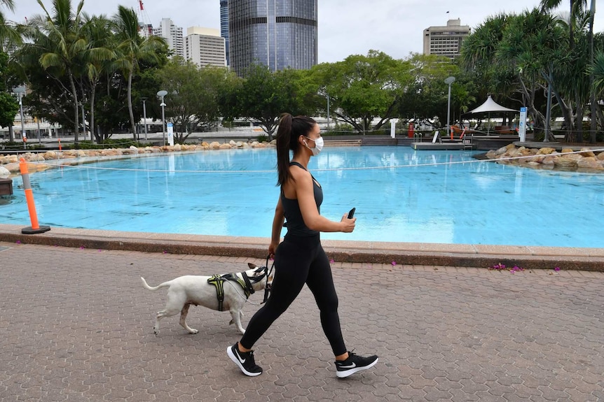 A woman walks a dog in a face mask near a pool.