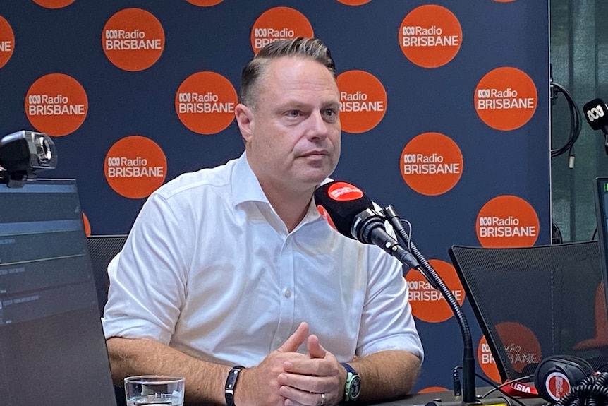 A man in a white business shirt sitting at a microphone. In the background is a wall of red circle logos that say Radio Brisbane