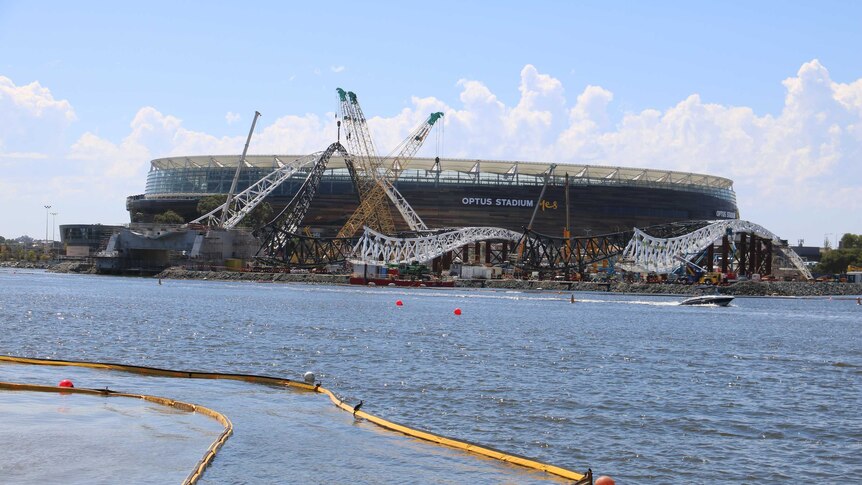 The Matagarup Bridge under construction alongside the Swan River with Perth Stadium behind it and water in the foreground.