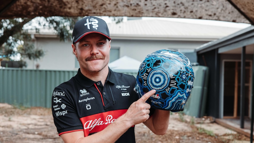 A moustachioed man in a dark cap points at a racing helmet he is holding. It features an intricate Indigenous design.