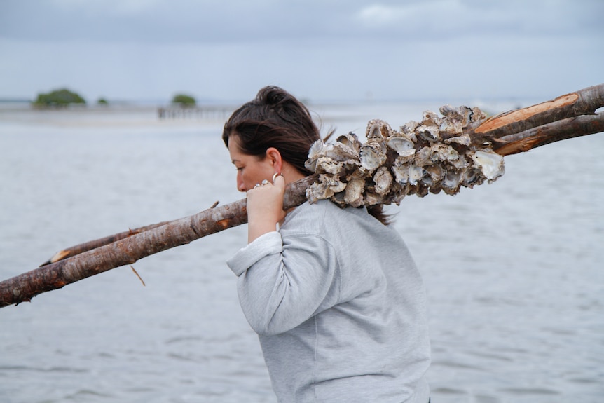 A brunette holds a wooden stake, onto which oyster shells are attached, over her shoulder.