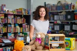 A woman with dark curly hair wearing a black and white striped tshirt stands surrounded by books and toys