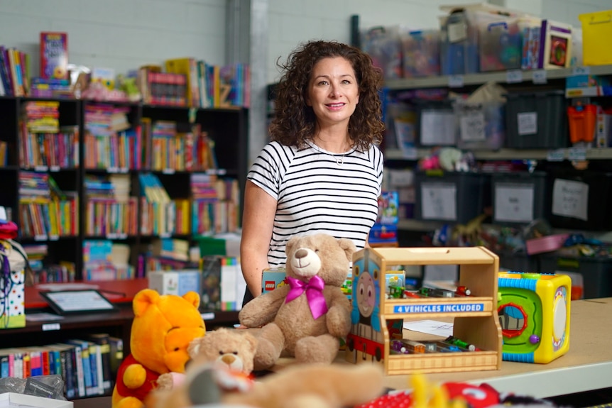 A woman with dark curly hair wearing a black and white striped tshirt stands surrounded by books and toys