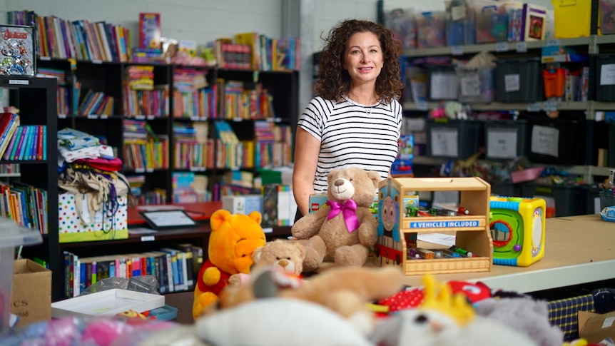 A woman with dark curly hair wearing a black and white striped tshirt stands surrounded by books and toys