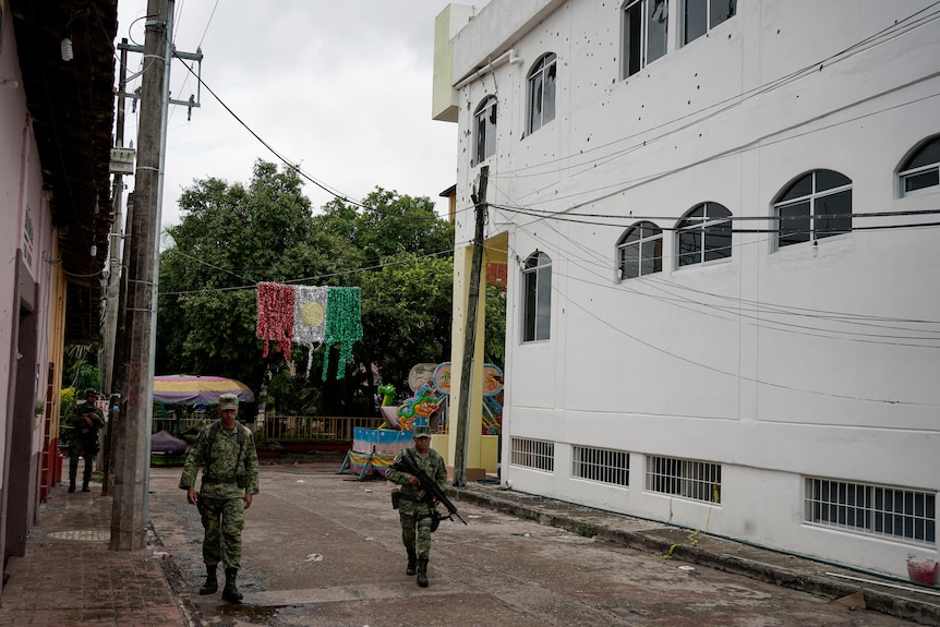two soldiers, one with a gun, on patrol, walk past a bullet-riddled two-storey building
