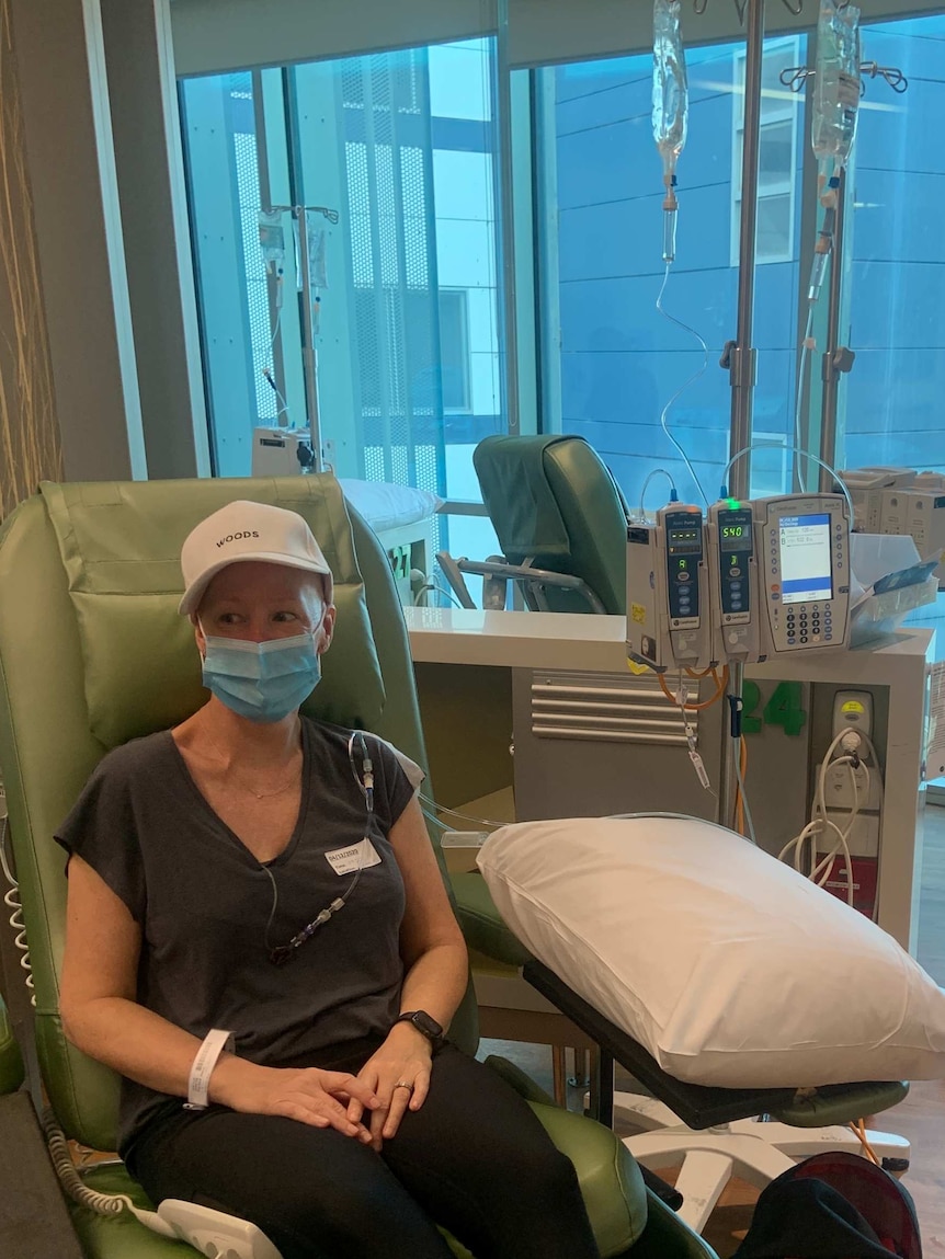 Jo sits on a green hospital chair, in front of a medical drip system. She is wearing a facemask and a white cap.