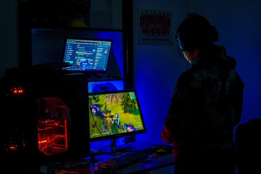 A teenage boy in a headset stands at two computer screens playing video games in a dark room.