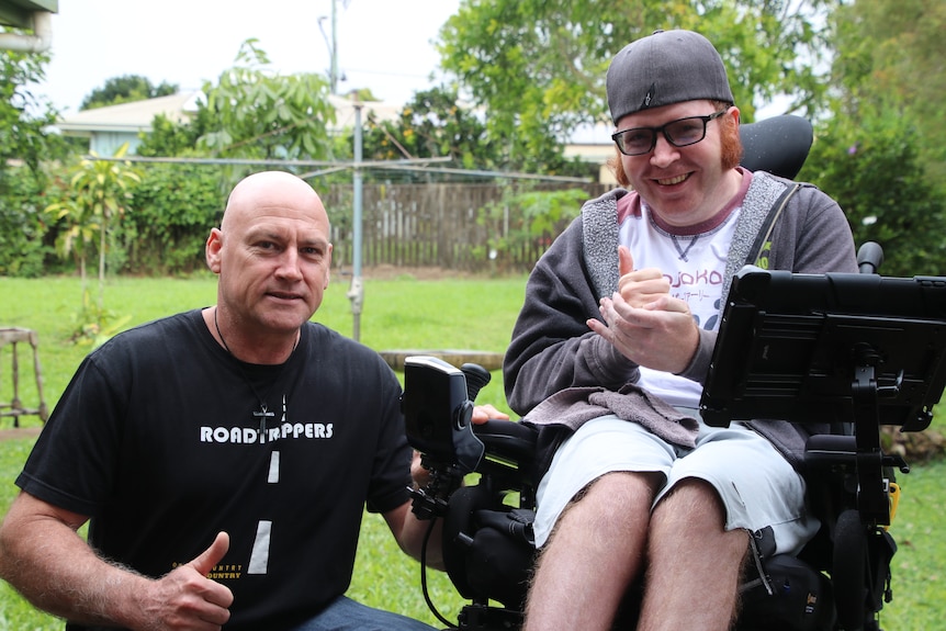 A young man wearing a cap sits in a wheelchair smiling with the his carer to his right 