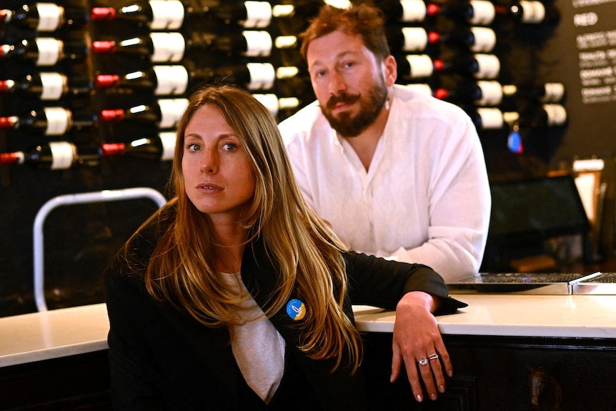 A white woman with long hair sits in front of a counter, which a white man behind her leans on. Wine bottles on wall behind him.