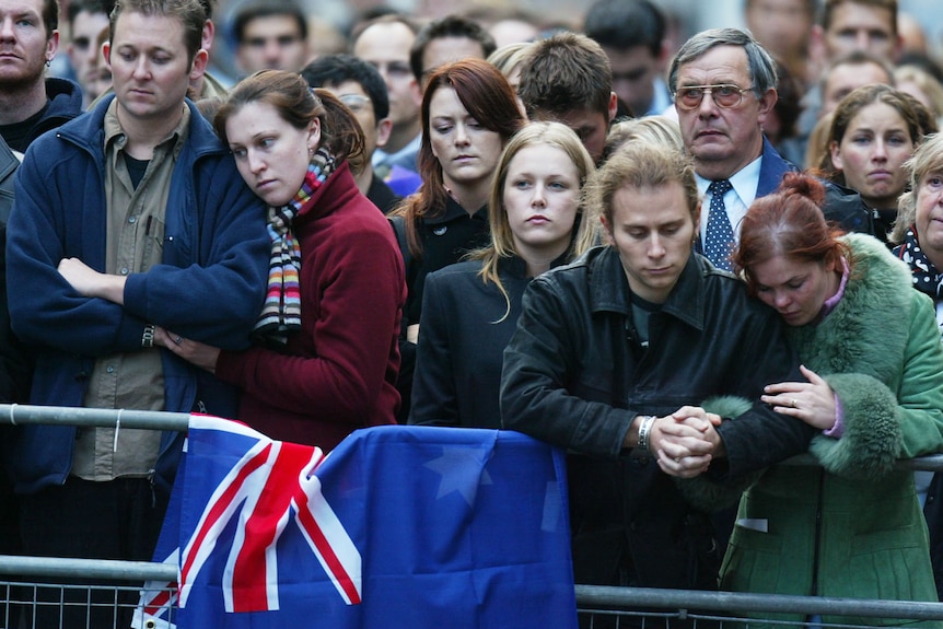 A group of solemn-faced people stand together behind a barricade draped with the Australian flag