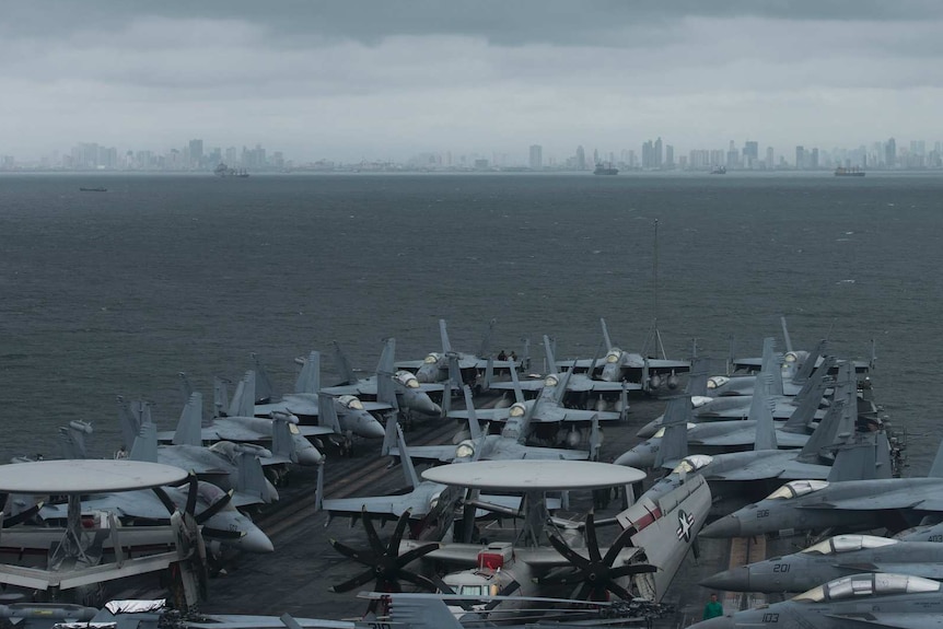 On an overcast day, you view an aircraft carrier stacked with US jets and helicopters as you view a distant Manila skyline.