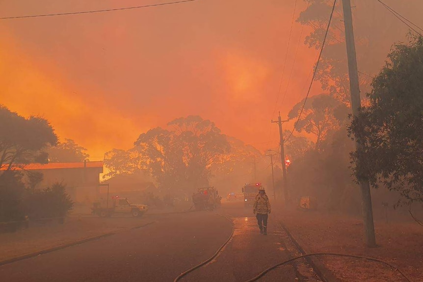 A firefighter walks along a street with a blood red sky and smoke behind him.