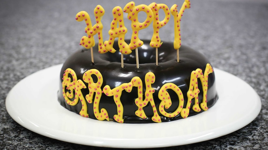 A round glazed chocolate cake with Happy Birthday letters sitting on a white plate on a slate kitchen top.