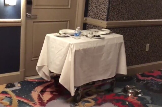 A room service cart in the hallway of a hotel has a camera under a dirty dish, with cord running down under hotel room door.
