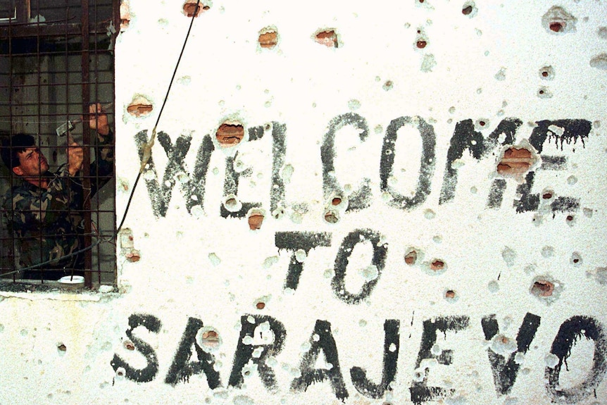 A soldier works in a damaged building riddled with bullet holes with a sign on the wall saying welcome to Sarajevo.