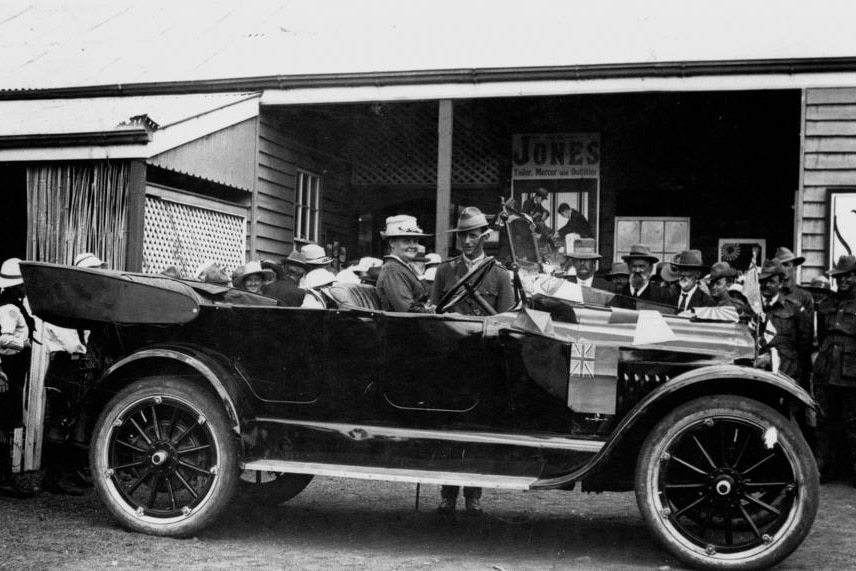 A woman sits in a 1920s car, people are standing in the background. Black and white picture. 