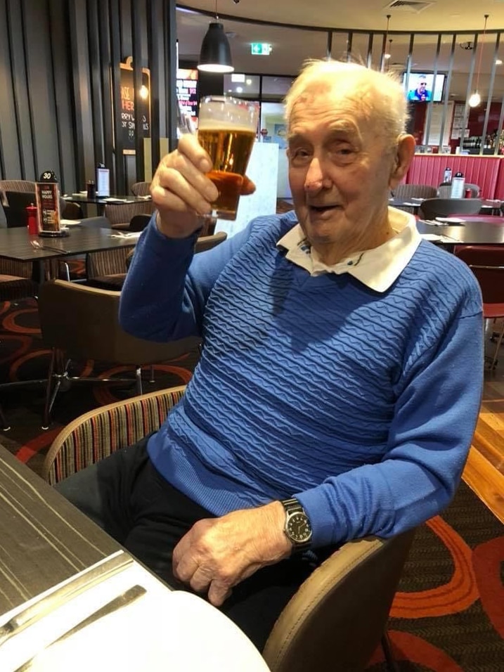 Alf Jordan wearing a blue jumper, smiles and playfully holds up a beer in a restaurant.