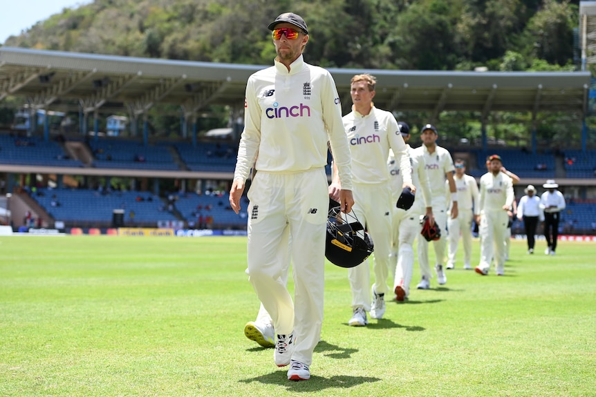 Joe Root walks in front of a group of men in cricket whites