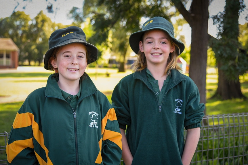 Two children in green school uniforms wearing green bucket hats smiling.