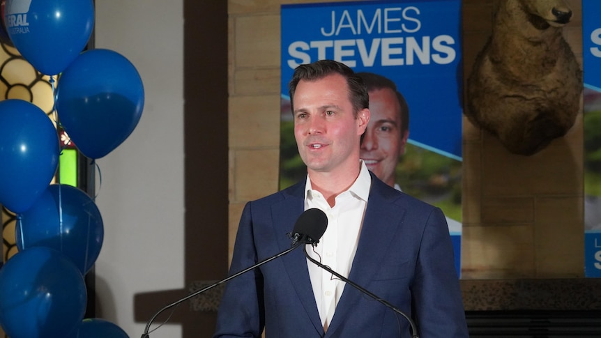 A man wearing a blue suit without a te  in front of a sign with his name and picture and blue balloons