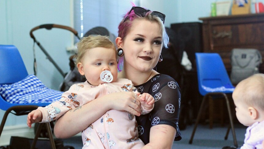 A young woman with multi-coloured hair is holding a year-old baby