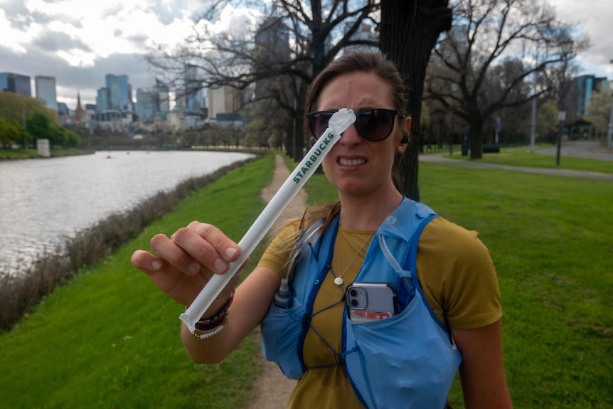 A woman holds a plastic straw close to the camera with the Yarra River in the background.