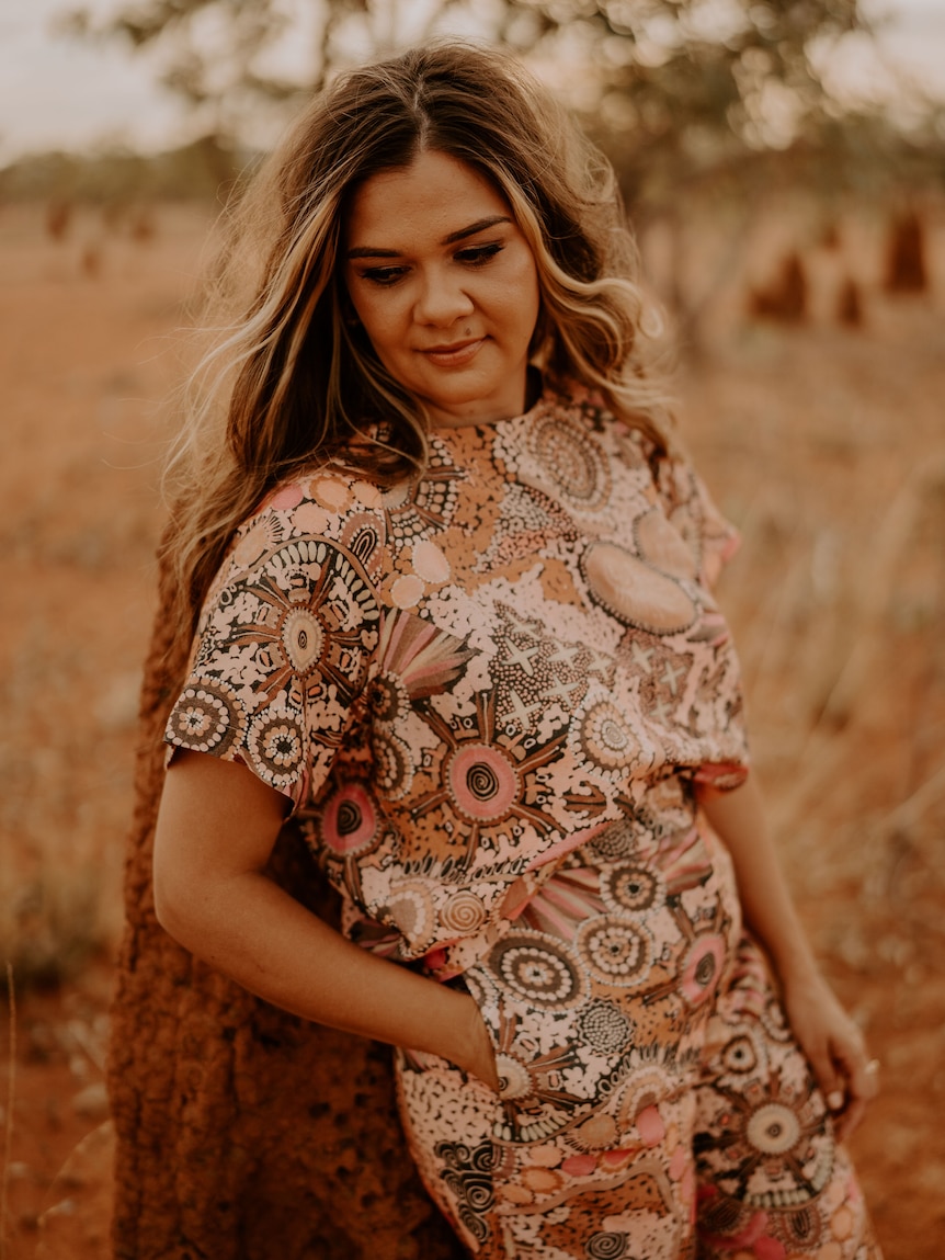An Indigenous model wears a shirt and pants in a pink, Indigenous artwork pattern, in a desert landscape